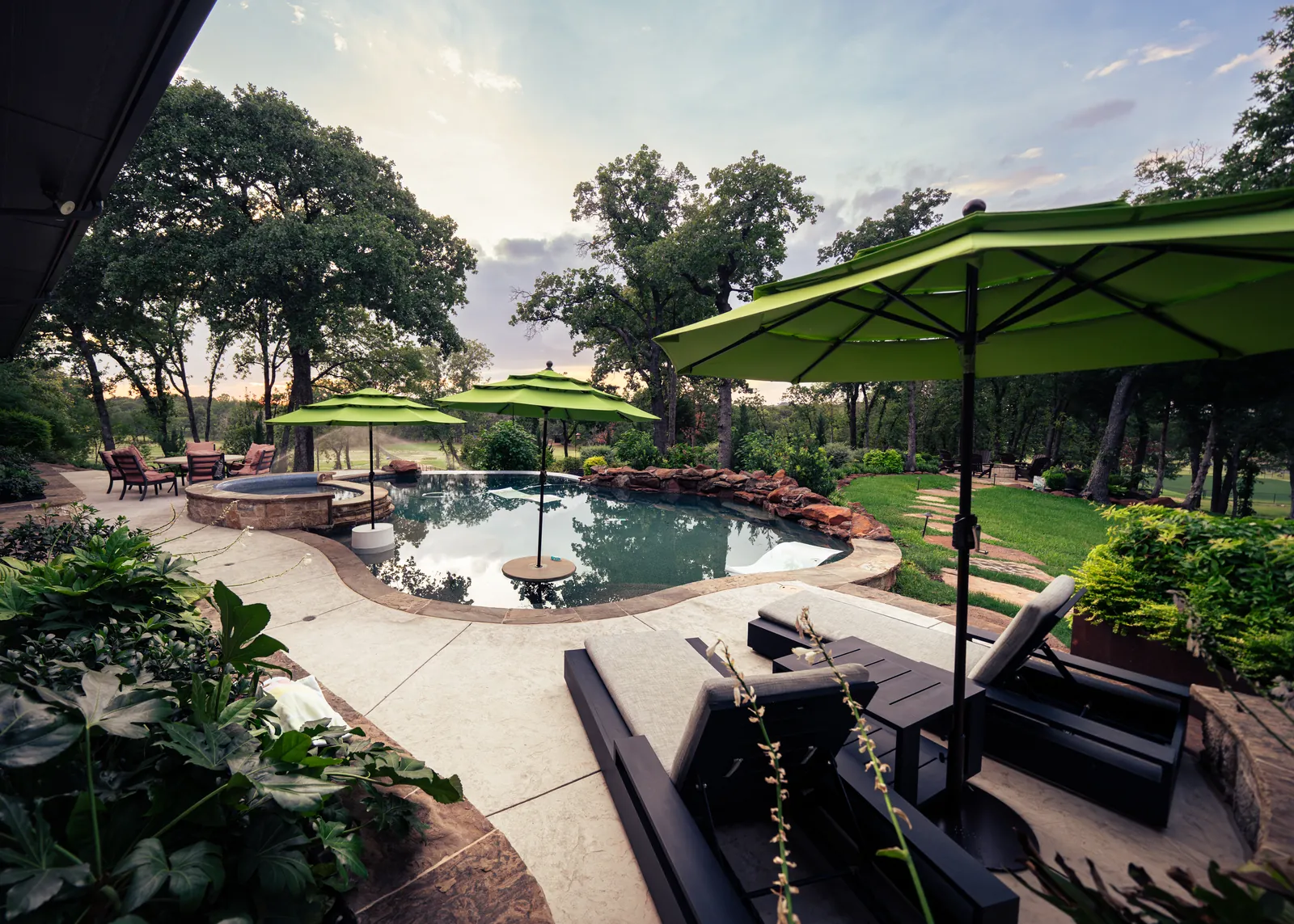Backyard pool and spa with stone patio, shade umbrellas, and landscaping at dusk by Greenview Landscaping in Fort Worth