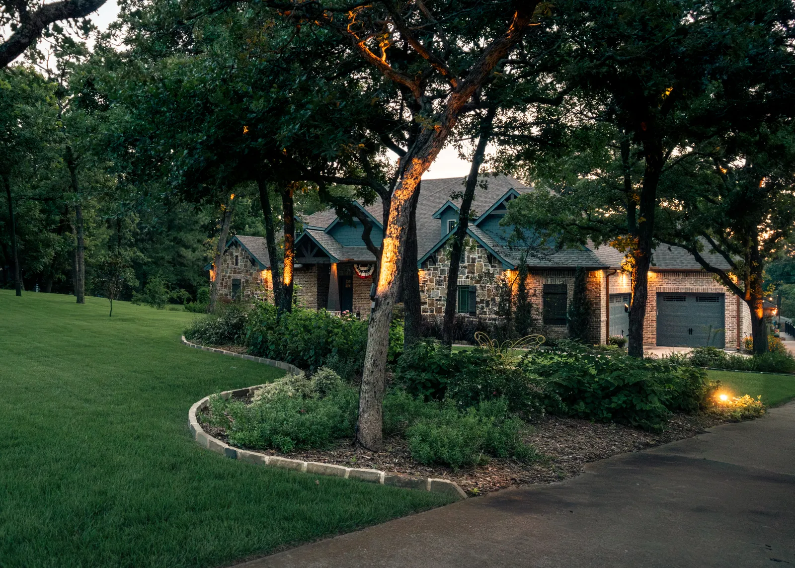 Stone home at dusk with landscape lighting, curved planting beds, and a manicured lawn by Greenview Landscaping in Fort Worth